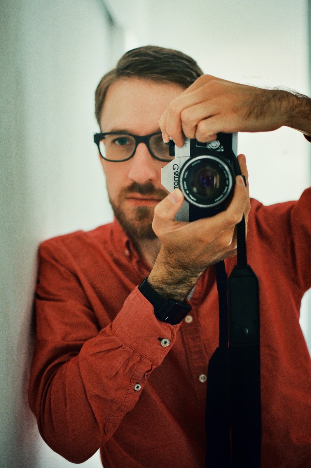 A close-up mirror selfie taken with a Canon FTb QL camera fitted with a 50 mm lens. I’m wearing a red shirt and a black wristwatch, with a black camera strap visible. The background is softly blurred, showing white walls lit by indirect sunlight.