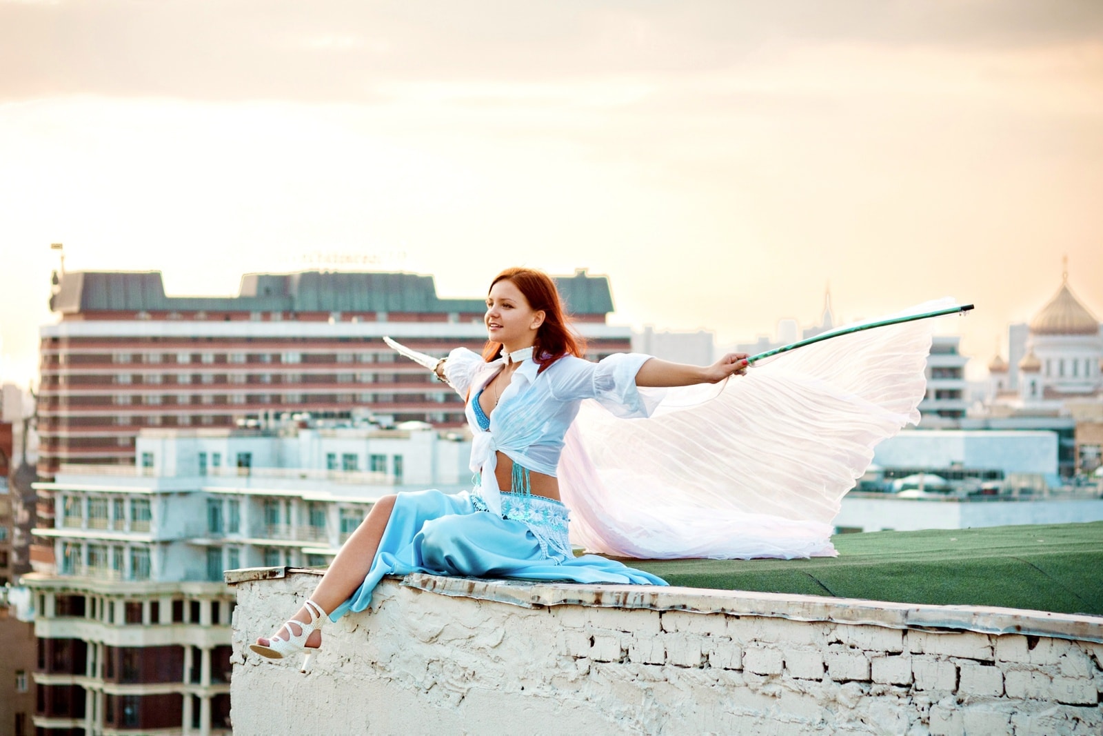 Beautiful girl is sitting on the edge of rooftop in Moscow. She looks somewhere ahead with a happy face. She has Isis wings. It is close to sunset, the sky is orangish. The Cathedral of Christ the Saviour is seen somewhat a kilometer away in the background.